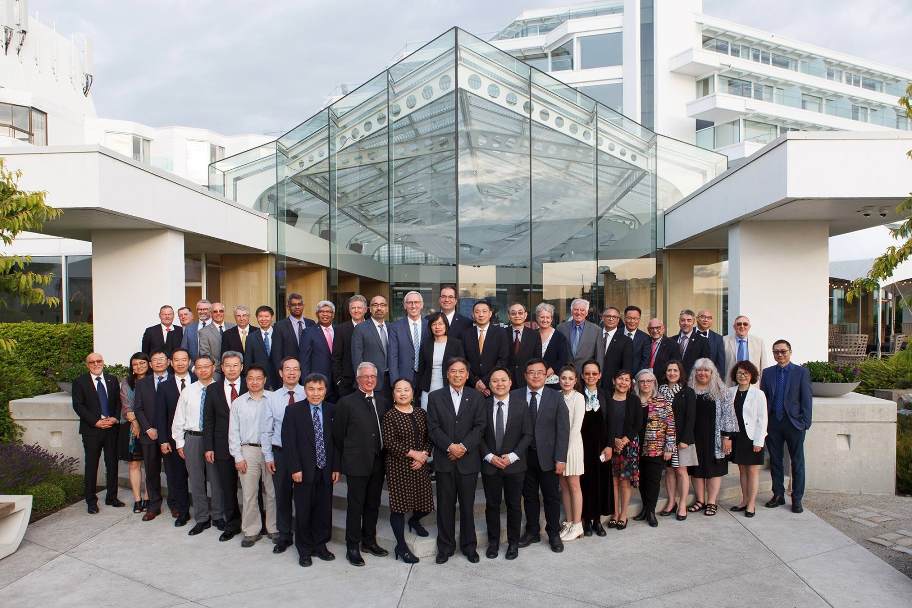 A large group of people, approximately 40 individuals from the CAE Fellows, pose for a group photo in front of The Inn at Laurel Point, a modern building with glass architecture. The group is diverse, consisting mostly of men with a few women, all dressed in formal business attire. They are arranged in several rows, with some standing on higher steps to be visible. The hotel behind them features a prominent glass facade and contemporary design.