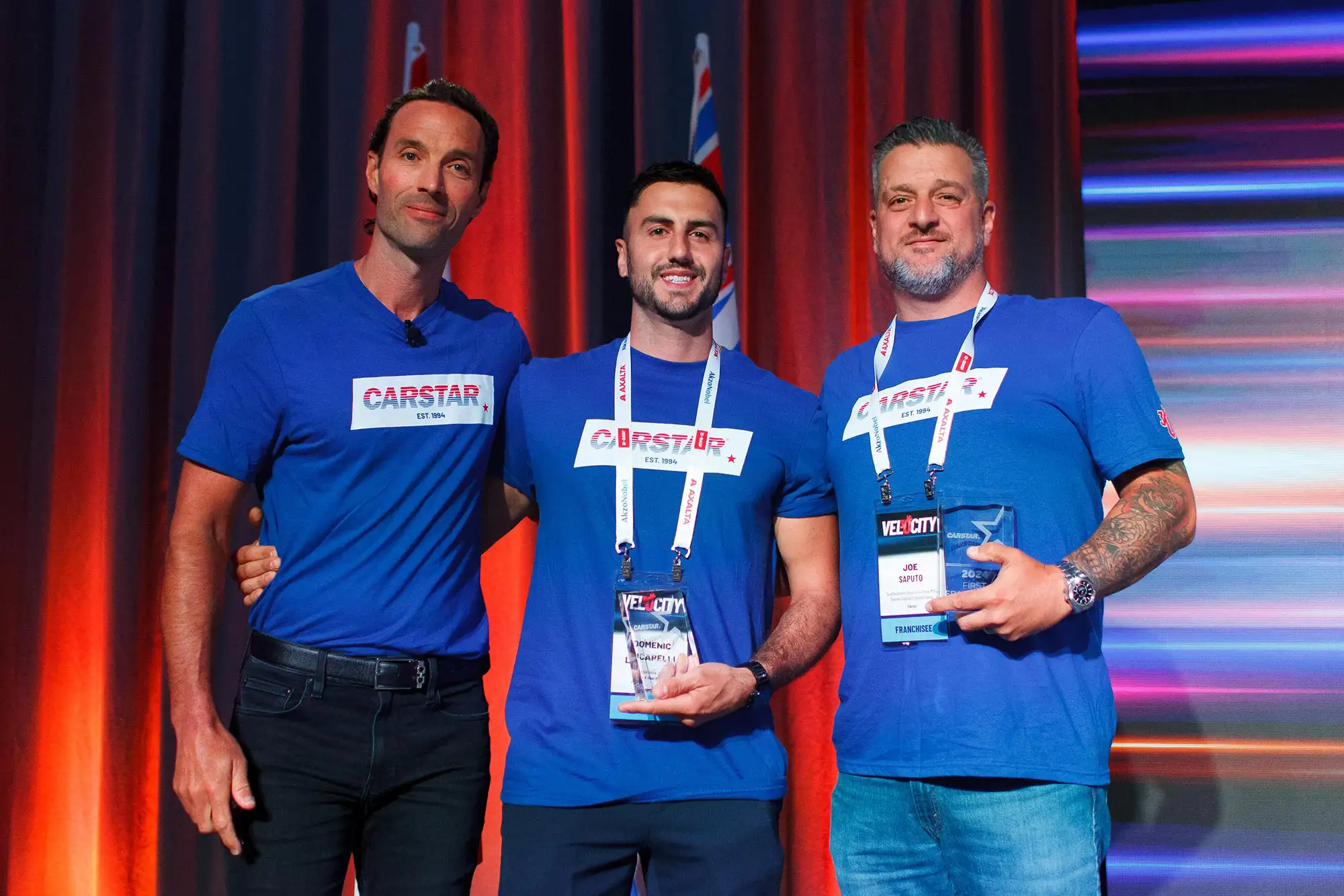 Three men in blue shirts stand on stage, one holding an award, smiling at the audience during corporate event in Victoria BC